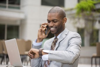 Closeup handsome young businessman working with laptop outdoors talking on mobile phone looking at his wristwatch. Time is money